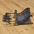 Traîneau de Noël sombre à motifs ajourés et patins recourbés, attelé à trois rennes, vu de trois quarts sur une table en bois.
