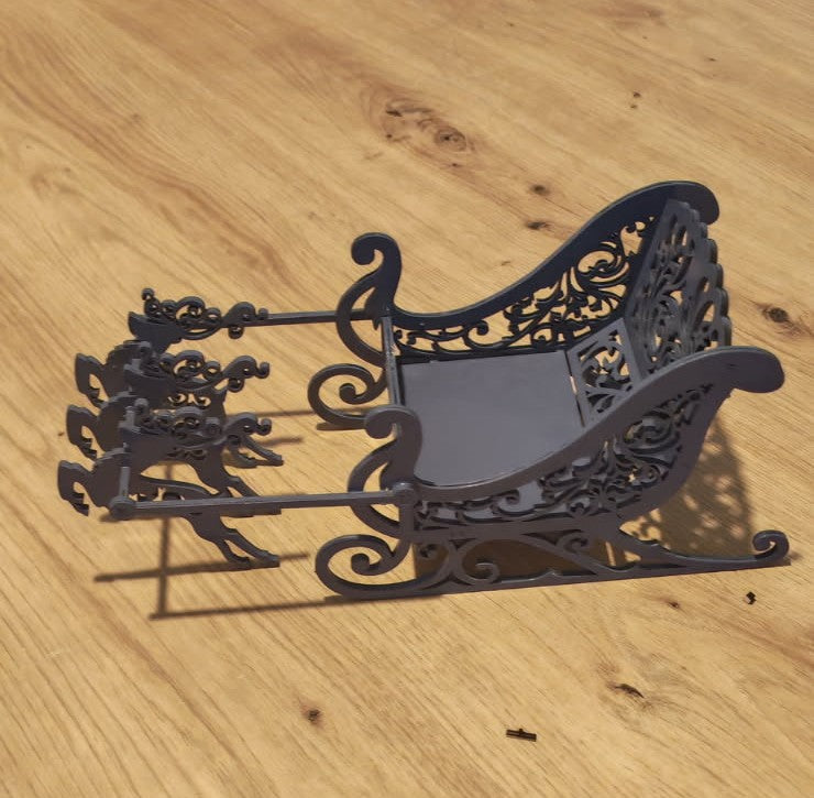 Traîneau de Noël sombre à motifs ajourés et patins recourbés, attelé à trois rennes, vu de trois quarts sur une table en bois.