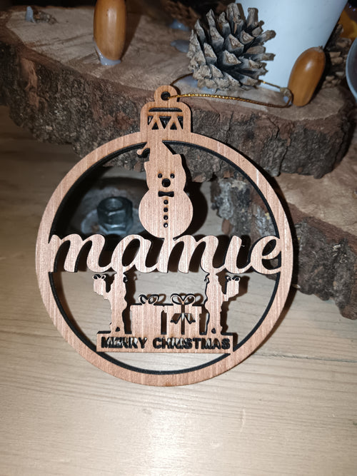 Boule de Noël en bois ajouré avec inscription Mamie, bonhomme de neige et cadeaux, posée devant des rondins