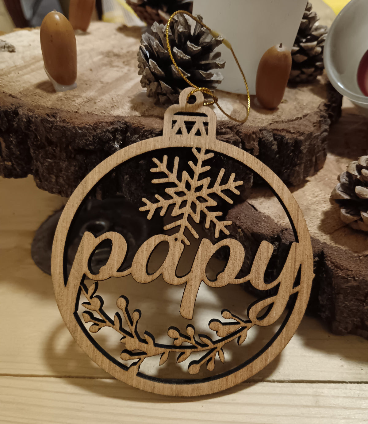 Boule de Noël en bois ajouré avec inscription Papy, flocon et branches décoratives, posée devant des rondins