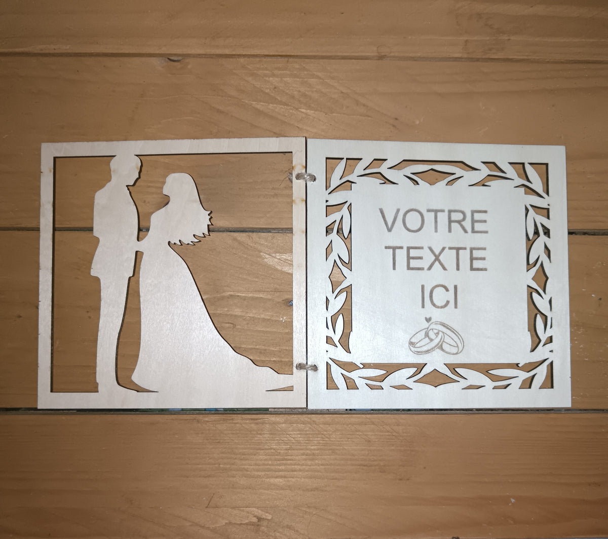 Faire-part de mariage en bois ouvert, avec silhouettes de mariés à gauche et espace de texte entouré de feuillage à droite