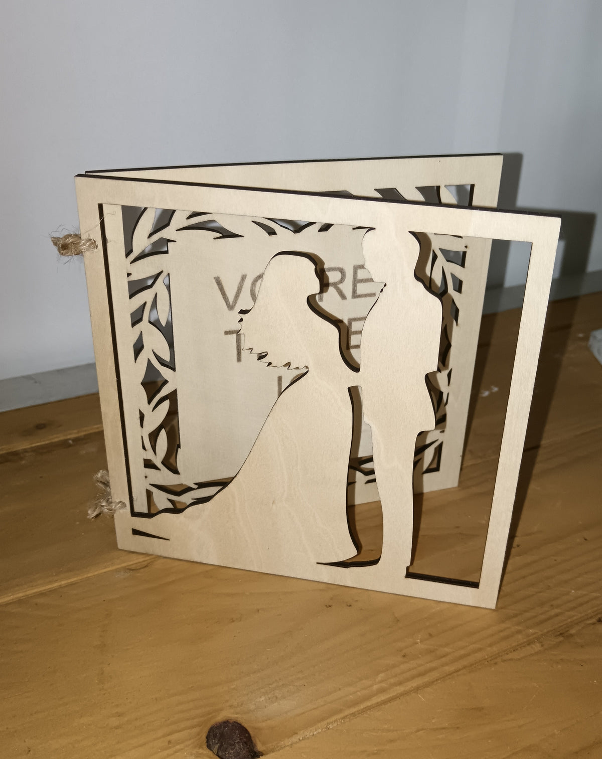 Faire-part de mariage en bois entrouvert, avec silhouettes de mariés découpées et bordure de feuillage