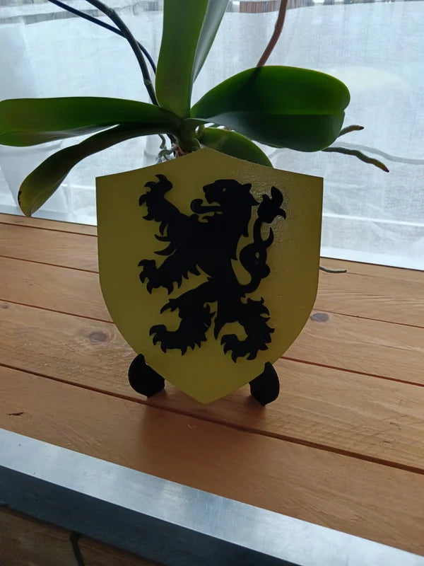 Petit blason en bois jaune avec Lion des Flandres noir, présenté sur un chevalet devant une plante