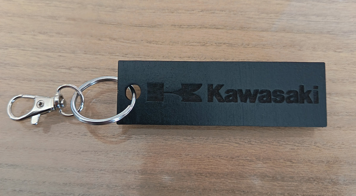 Porte-clés rectangulaire en bois noir avec logo Kawasaki et attache métallique, posé sur une surface en bois clair