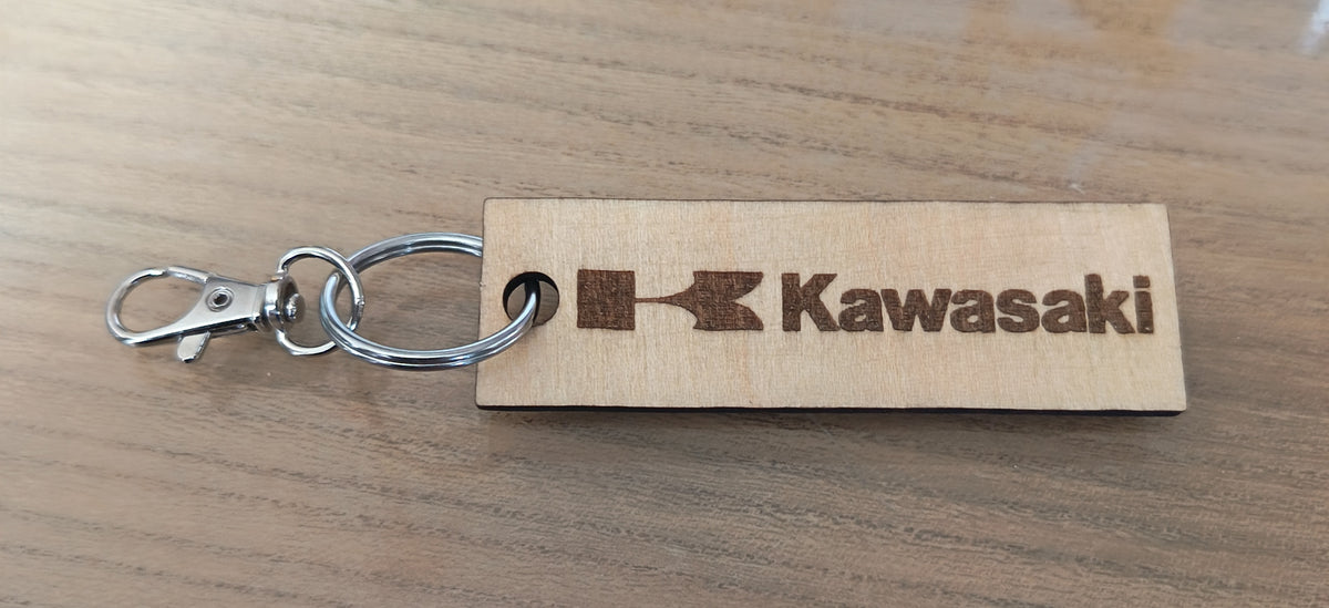Porte-clés rectangulaire en bois clair avec logo Kawasaki et attache métallique, posé sur une surface en bois